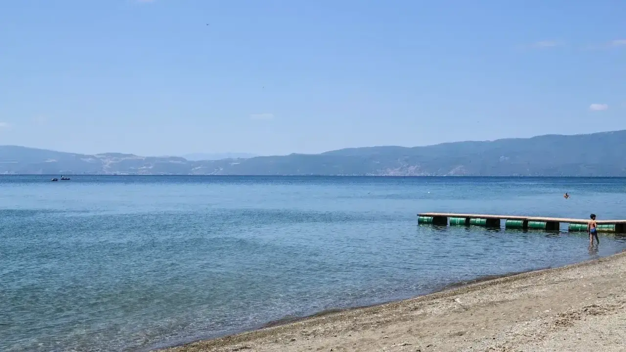 Plaże nad Jeziorem Ochrydzkim. Dziecko wchodzi do wody z pomostu, w tle góry i błękitne niebo.