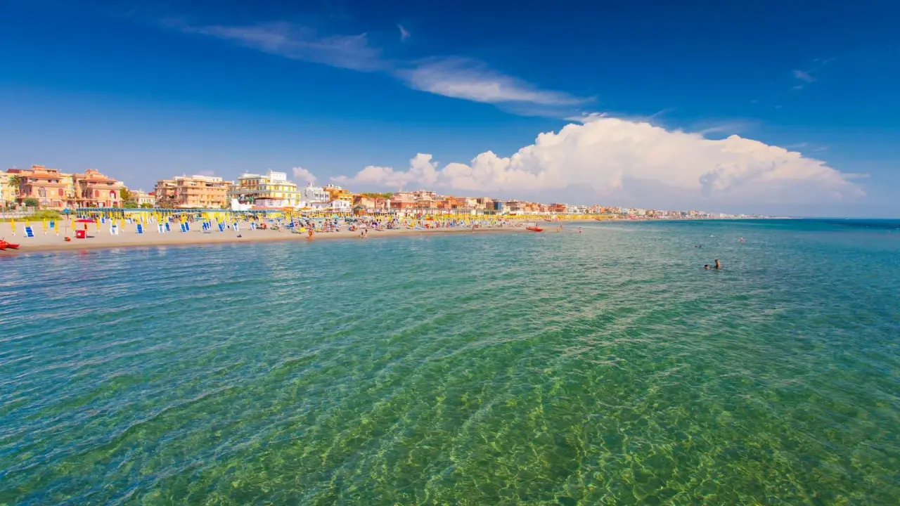 Słoneczna plaża w Fiumicino z kolorowymi parasolami, budynkami w tle i turkusową wodą, w której pływają ludzie.
