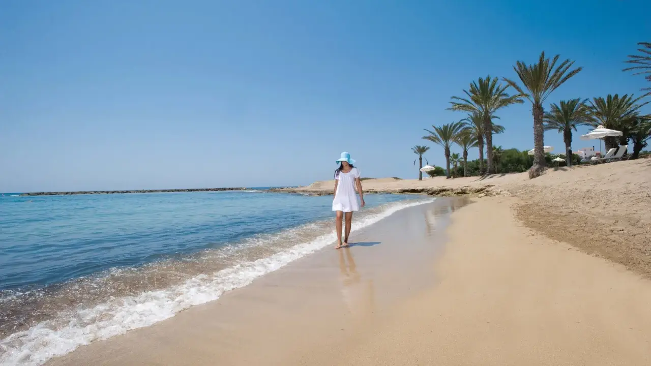 Dziewczyna w białej sukience i niebieskim kapeluszu spaceruje po piaszczystej plaży na Cyprze. Widok idealny, by zobaczyć, co oferuje wyspa.