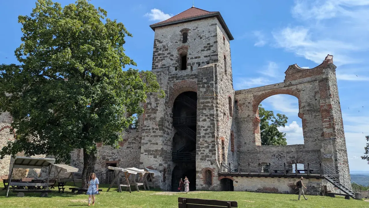 Ruiny zamku Tenczyn, z wieżą i łukiem, na tle błękitnego nieba. Widać drzewo i ludzi spacerujących po trawniku.