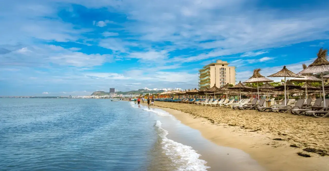 Słoneczna plaża w Golem, Albania, z rzędem leżaków i parasoli. W oddali widać hotel i miasto.