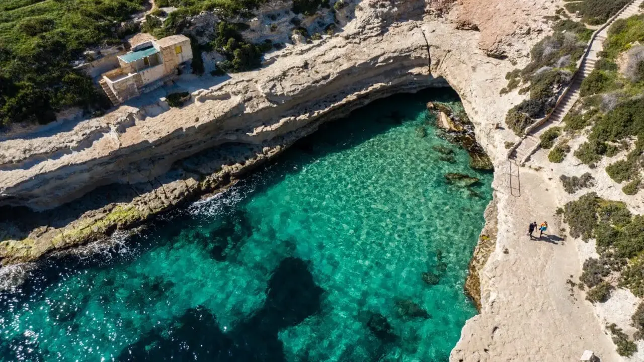 Malta plaże: turkusowa woda w zatoczce, skaliste wybrzeże i mały domek na klifie. Dwie osoby spacerują ścieżką.