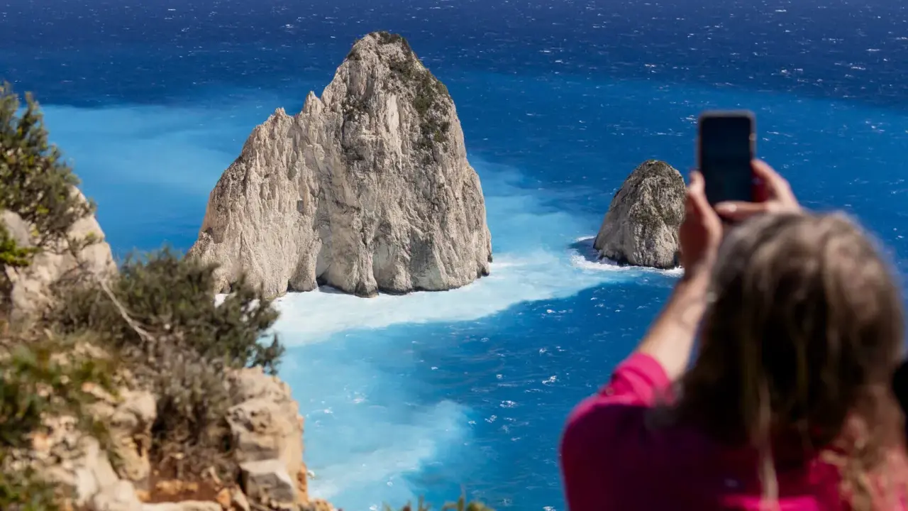Widok na turkusowe morze i skaliste wysepki. Kobieta robi zdjęcie telefonem. Czy Zakynthos warto odwiedzić? Tak!