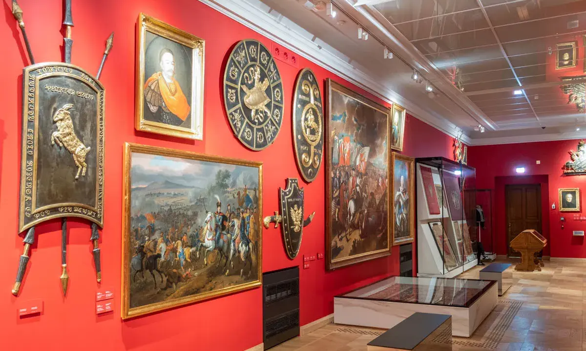 Wnętrze jednego z muzeów w Krakowie. Czerwone ściany zdobią obrazy batalistyczne, portrety i historyczne tarcze.