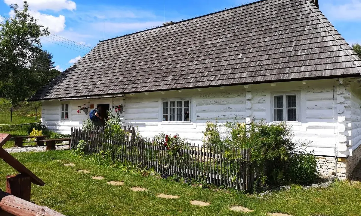 Tradycyjna chata kocjana z drewnianym dachem i białymi ścianami, otoczona zielenią i kamiennym płotem.