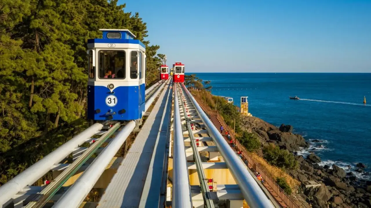 Kolejka linowa wzdłuż wybrzeża, z widokiem na turkusowe morze i zielone drzewa. To widok z **Jeju, Korea**.