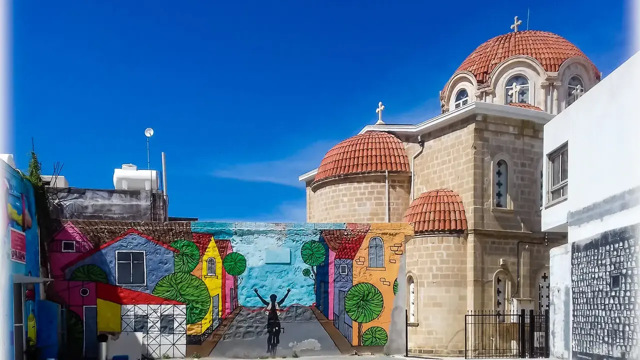 Kolorowy mural z rowerzystą i domkami na tle kościoła z kopułami. Pafos Cypr centrum.