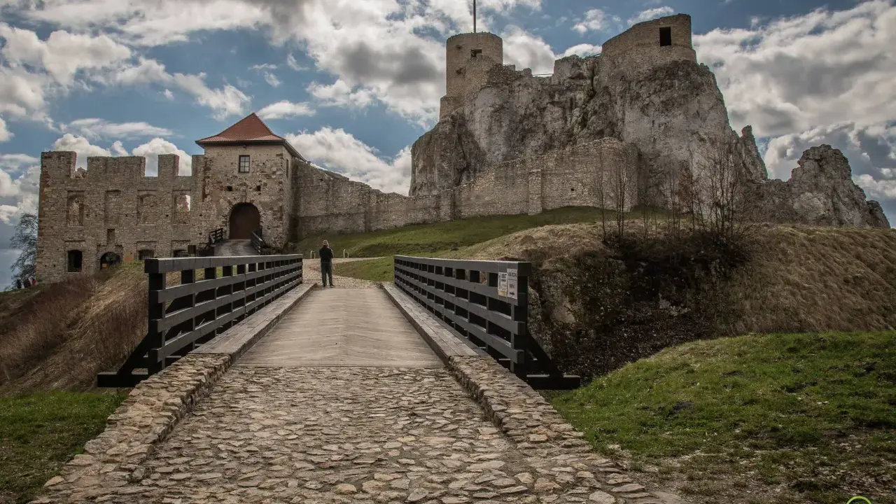Kamienny most prowadzi do ruin zamku w Olkuszu. Na szczycie skalistego wzgórza widać wieżę z polską flagą.