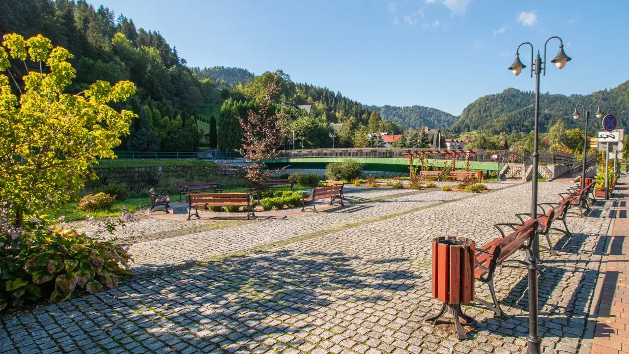 Słoneczna promenada Szczawnica z kamienną nawierzchnią, ławkami i latarniami, otoczona zielenią i górami.