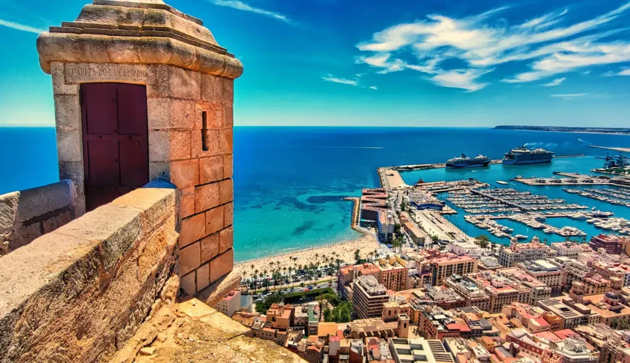 Alicante co zobaczyć: widok z zamku na port, plażę i miasto. Widać wieżyczkę strażniczą, jachty i statki wycieczkowe.