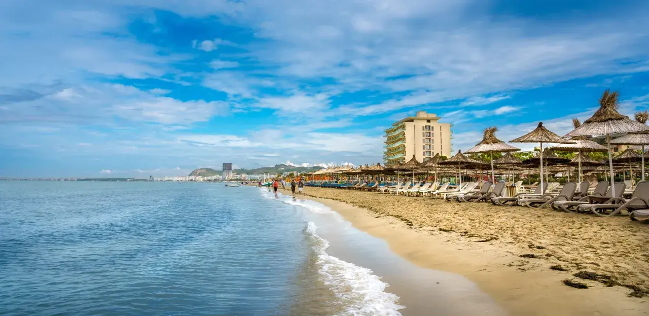 Słoneczna plaża w Durrës, Albania, z rzędami leżaków i parasoli. W tle widać hotel i miasto, a na pierwszym planie falujące morze. Idealne miejsce na wakacje i odkrywanie atrakcji.