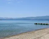 Plaże nad Jeziorem Ochrydzkim. Dziecko wchodzi do wody z pomostu, w tle góry i błękitne niebo.
