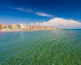 Słoneczna plaża w Fiumicino z kolorowymi parasolami, budynkami w tle i turkusową wodą, w której pływają ludzie.