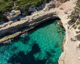 Malta plaże: turkusowa woda w zatoczce, skaliste wybrzeże i mały domek na klifie. Dwie osoby spacerują ścieżką.