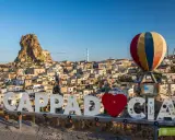 Cappadocia, Turcja zwiedzanie na własną rękę. Balon i napis "Cappadocia" na tle skalnych domów i twierdzy.