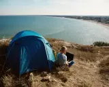 Niebieski namiot i osoba z laptopem na wzgórzu, z widokiem na lazurowe wybrzeże. Idealne miejsce na camping.