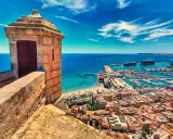 Alicante co zobaczyć: widok z zamku na port, plażę i miasto. Widać wieżyczkę strażniczą, jachty i statki wycieczkowe.