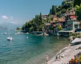 Urokliwa plaża w Bergamo, z kolorowymi domkami na wzgórzu, łodziami na wodzie i dziećmi bawiącymi się nad brzegiem.