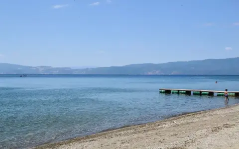 Plaże nad Jeziorem Ochrydzkim. Dziecko wchodzi do wody z pomostu, w tle góry i błękitne niebo.