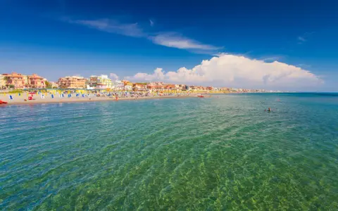Słoneczna plaża w Fiumicino z kolorowymi parasolami, budynkami w tle i turkusową wodą, w której pływają ludzie.