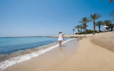 Dziewczyna w białej sukience i niebieskim kapeluszu spaceruje po piaszczystej plaży na Cyprze. Widok idealny, by zobaczyć, co oferuje wyspa.
