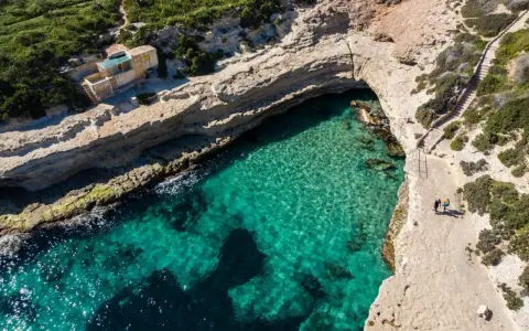 Malta plaże: turkusowa woda w zatoczce, skaliste wybrzeże i mały domek na klifie. Dwie osoby spacerują ścieżką.