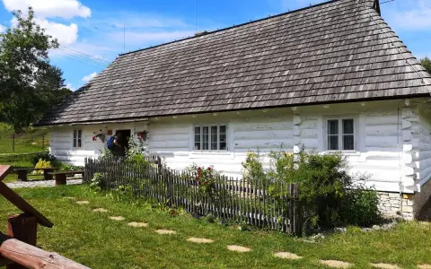 Tradycyjna chata kocjana z drewnianym dachem i białymi ścianami, otoczona zielenią i kamiennym płotem.