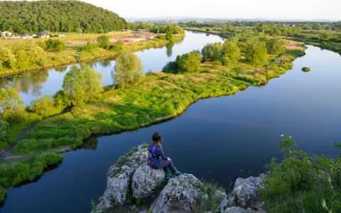 Dziewczyna siedzi na skale, podziwiając malowniczy krajobraz Bielańsko-Tynieckiego Parku Krajobrazowego. Rzeka meandruje wśród zielonych wzgórz i pól.