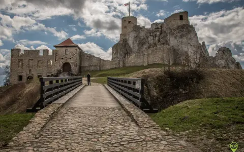 Kamienny most prowadzi do ruin zamku w Olkuszu. Na szczycie skalistego wzgórza widać wieżę z polską flagą.