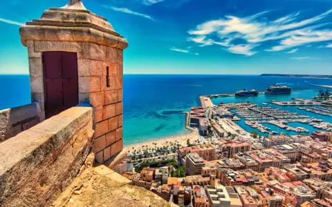 Alicante co zobaczyć: widok z zamku na port, plażę i miasto. Widać wieżyczkę strażniczą, jachty i statki wycieczkowe.