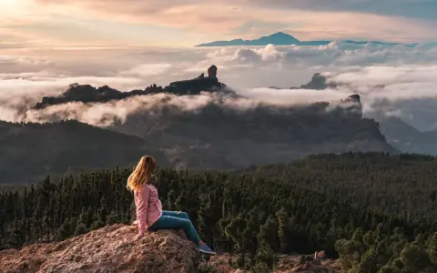 Kobieta siedzi na skale, podziwiając mgły i góry na Gran Canarii. W oddali widać Roque Nublo.