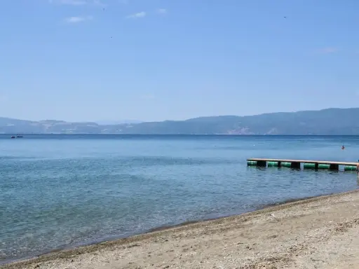 Plaże nad Jeziorem Ochrydzkim. Dziecko wchodzi do wody z pomostu, w tle góry i błękitne niebo.