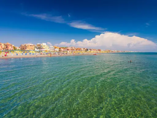 Słoneczna plaża w Fiumicino z kolorowymi parasolami, budynkami w tle i turkusową wodą, w której pływają ludzie.