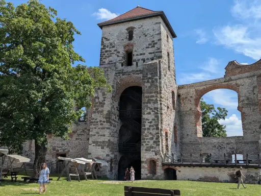 Ruiny zamku Tenczyn, z wieżą i łukiem, na tle błękitnego nieba. Widać drzewo i ludzi spacerujących po trawniku.