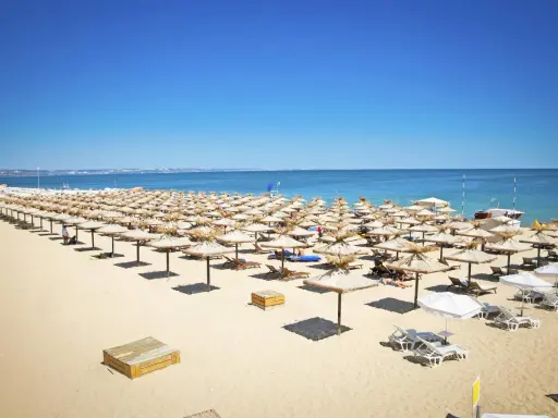 Słoneczny brzeg Bułgaria: Rzędy słomianych parasoli i leżaków na piaszczystej plaży, z błękitnym morzem w tle.