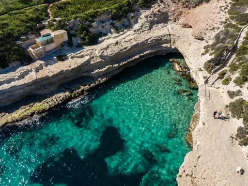 Malta plaże: turkusowa woda w zatoczce, skaliste wybrzeże i mały domek na klifie. Dwie osoby spacerują ścieżką.