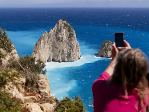 Widok na turkusowe morze i skaliste wysepki. Kobieta robi zdjęcie telefonem. Czy Zakynthos warto odwiedzić? Tak!