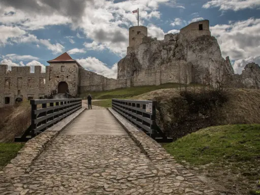 Kamienny most prowadzi do ruin zamku w Olkuszu. Na szczycie skalistego wzgórza widać wieżę z polską flagą.