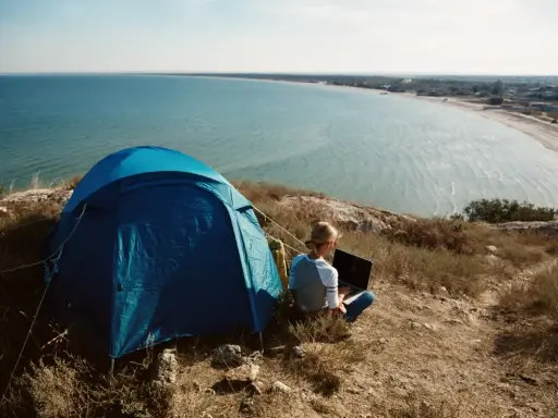 Niebieski namiot i osoba z laptopem na wzgórzu, z widokiem na lazurowe wybrzeże. Idealne miejsce na camping.