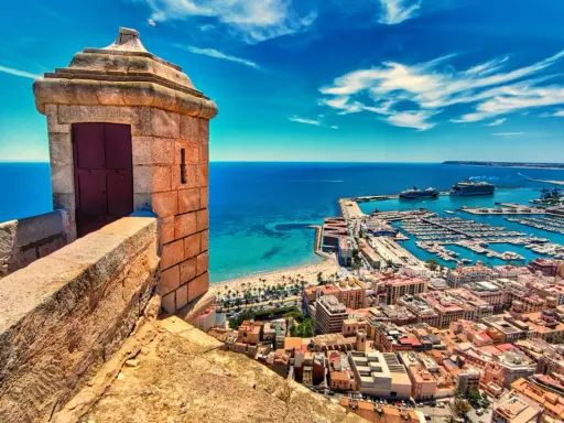 Alicante co zobaczyć: widok z zamku na port, plażę i miasto. Widać wieżyczkę strażniczą, jachty i statki wycieczkowe.