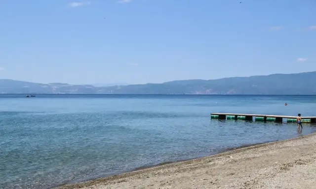 Plaże nad Jeziorem Ochrydzkim. Dziecko wchodzi do wody z pomostu, w tle góry i błękitne niebo.