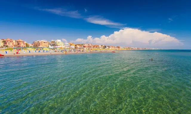 Słoneczna plaża w Fiumicino z kolorowymi parasolami, budynkami w tle i turkusową wodą, w której pływają ludzie.