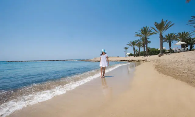 Dziewczyna w białej sukience i niebieskim kapeluszu spaceruje po piaszczystej plaży na Cyprze. Widok idealny, by zobaczyć, co oferuje wyspa.