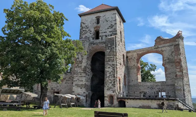 Ruiny zamku Tenczyn, z wieżą i łukiem, na tle błękitnego nieba. Widać drzewo i ludzi spacerujących po trawniku.