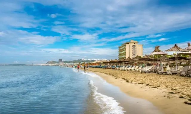 Słoneczna plaża w Golem, Albania, z rzędem leżaków i parasoli. W oddali widać hotel i miasto.