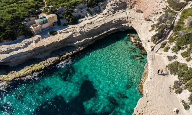 Malta plaże: turkusowa woda w zatoczce, skaliste wybrzeże i mały domek na klifie. Dwie osoby spacerują ścieżką.