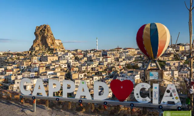 Cappadocia, Turcja zwiedzanie na własną rękę. Balon i napis "Cappadocia" na tle skalnych domów i twierdzy.
