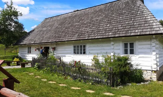 Tradycyjna chata kocjana z drewnianym dachem i białymi ścianami, otoczona zielenią i kamiennym płotem.