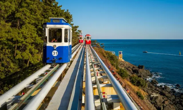 Kolejka linowa wzdłuż wybrzeża, z widokiem na turkusowe morze i zielone drzewa. To widok z **Jeju, Korea**.