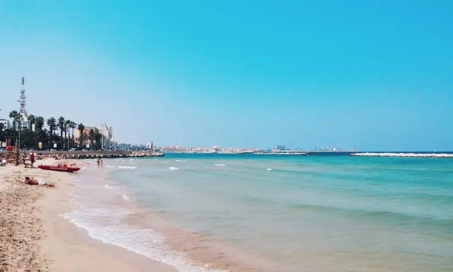 Słoneczna bari plaża z turkusową wodą i palmami w tle. Czerwona łódź ratownicza na piasku.